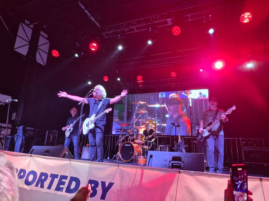 Singer with arms stretched wide at centre stage under red stage lights, bandmates on either side