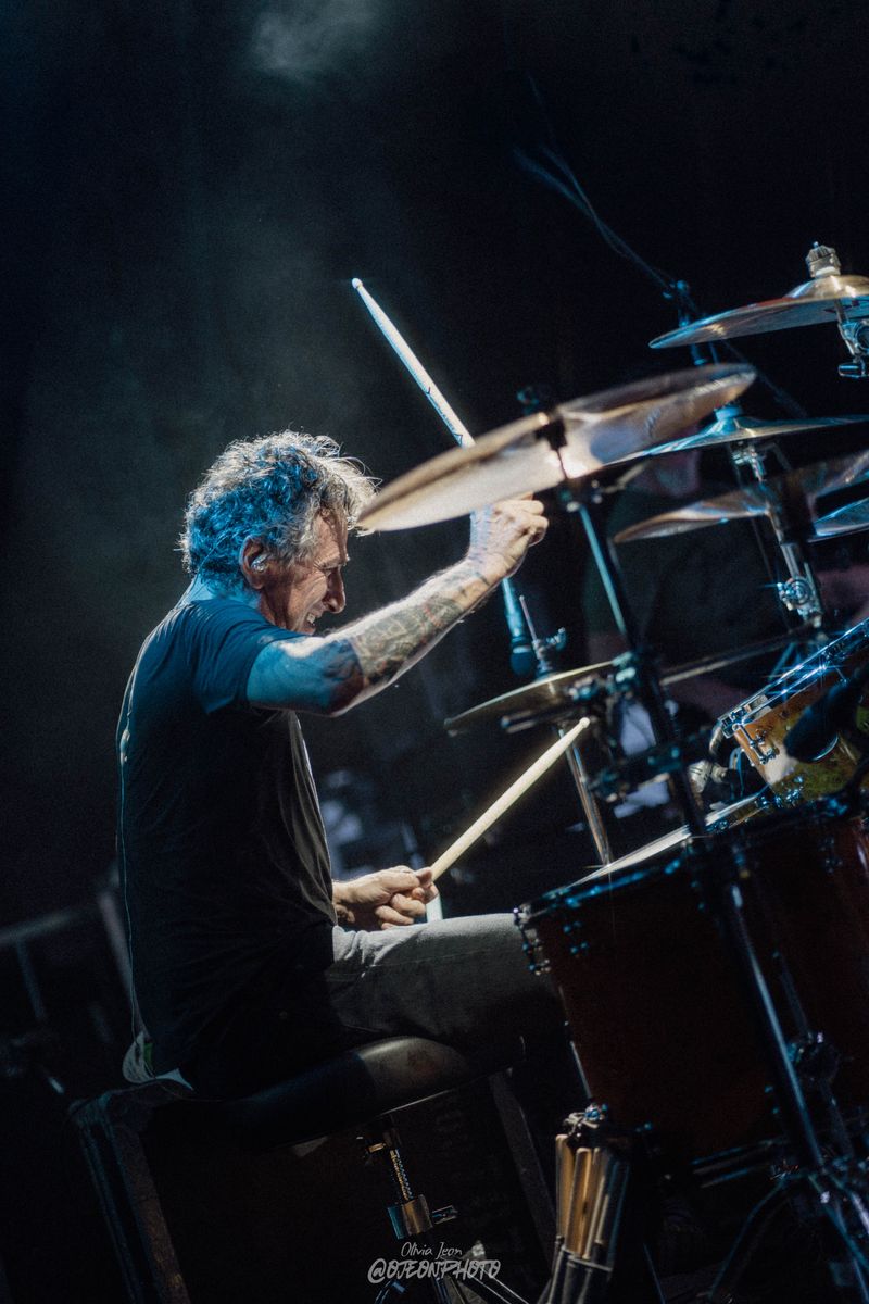 Drummer mid-hit with sticks raised, grinning, lit by blue haze