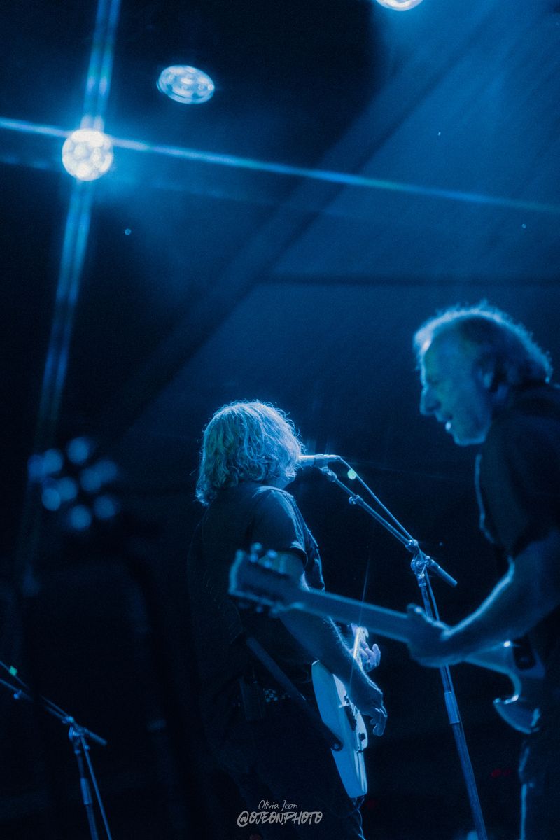 Two guitarists in deep blue stage light, one singing at the mic while the other leans in