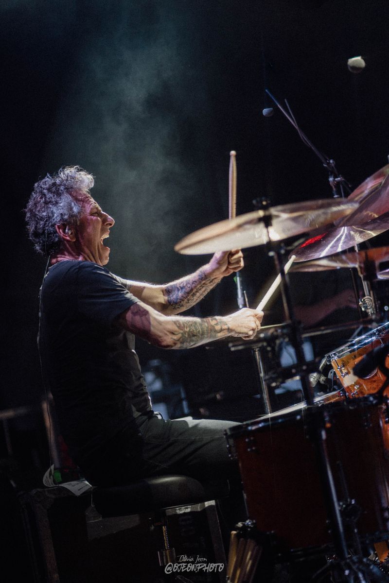 Drummer shouting while playing, cymbals ringing and smoke drifting through the stage light